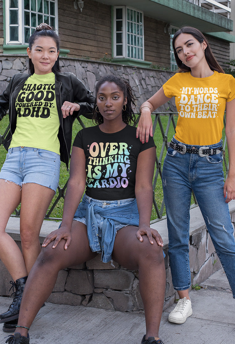 Three women wearing colorful t-shirts with text, standing outdoors against a wooden building.